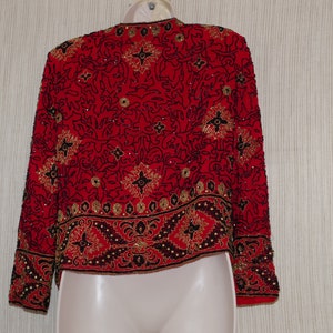 May include: A red, beaded jacket with intricate black and gold patterns. The jacket features long sleeves and a cropped design. The embellishments include sequins and beads, creating a decorative, formal look. The jacket is displayed on a mannequin.