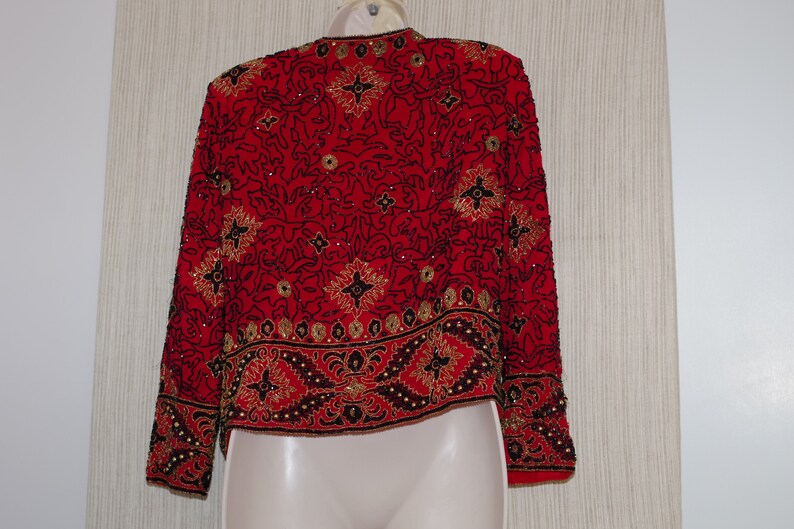 May include: A red, beaded jacket with intricate black and gold patterns. The jacket features long sleeves and a cropped design. The embellishments include sequins and beads, creating a decorative, formal look. The jacket is displayed on a mannequin.