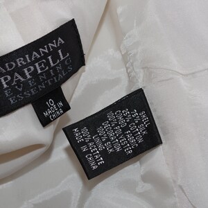 May include: Close-up of a white garment with two black tags. The tags read "ADRIANNA PAPELL EVENING ESSENTIALS" and include size 10. The second tag lists fabric content: 75% silk, 25% cotton, and 100% polyester.
