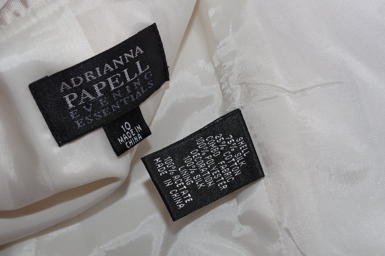 May include: Close-up of a white garment with two black tags. The tags read "ADRIANNA PAPELL EVENING ESSENTIALS" and include size 10. The second tag lists fabric content: 75% silk, 25% cotton, and 100% polyester.
