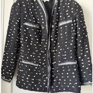 May include: A black tweed jacket with white and gray flecks, featuring a gray trim along the collar, pockets, and cuffs. The jacket has multiple silver buttons and two front pockets. The label reads "TERI JON".