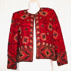 May include: A red jacket with intricate gold and black beadwork. The jacket features a round neckline and long sleeves. The design includes floral and geometric patterns, with a border of beads along the hem and cuffs. The jacket is displayed on a mannequin.