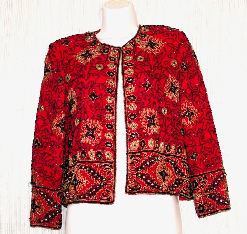 May include: A red jacket with intricate gold and black beadwork. The jacket features a round neckline and long sleeves. The design includes floral and geometric patterns, with a border of beads along the hem and cuffs. The jacket is displayed on a mannequin.