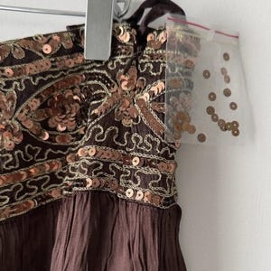 May include: A brown dress with copper-coloured sequins and embroidery. The dress is hanging on a hanger. A small plastic bag of sequins is attached to the hanger.
