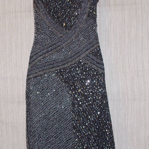 May include: A dark grey, knee-length dress with spaghetti straps. The dress is heavily embellished with sequins and beads, creating a shimmering effect. The bodice features a crisscross design, and the skirt has a straight silhouette. The dress is displayed on a mannequin.