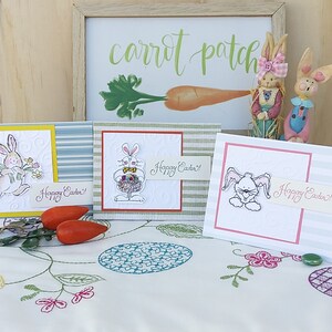 May include: Three handmade Easter cards with bunny illustrations and the text "Happy Easter!" on each card. The cards are on a white tablecloth with a green plant and two red carrots.