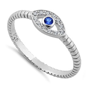 May include: A silver ring featuring an eye-shaped design. The ring has a textured band and a central eye motif with a blue center stone surrounded by small, clear stones. The ring is made of sterling silver, as indicated by the "925" stamp.