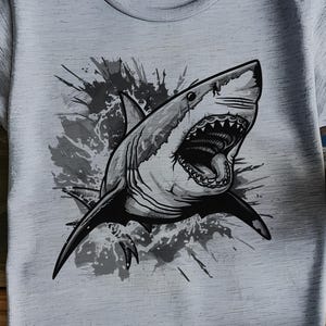 May include: A light gray t-shirt featuring a detailed black and white illustration of a shark with its mouth open. The shark is depicted with sharp teeth and is set against a splash-like background.