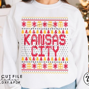 Puede incluir: Sudadera blanca con un diseño pixelado rojo y amarillo que dice "Kansas City". El diseño está rodeado de un patrón de copos de nieve y árboles de Navidad.