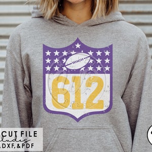 Könnte beinhalten: Ein verwittertes Design in Lila und Gelb mit der Nummer 612 und einem Fußball in einem Schild mit weißen Sternen. Das Design befindet sich auf einem grauen Hoodie.