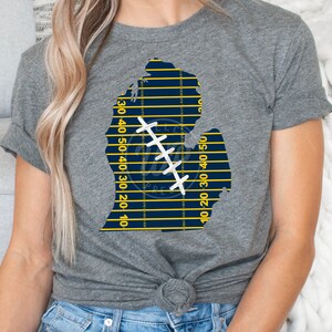 May include: A gray t-shirt with a blue and yellow outline of the state of Michigan. The outline is filled with a football field pattern and a white stitched football.