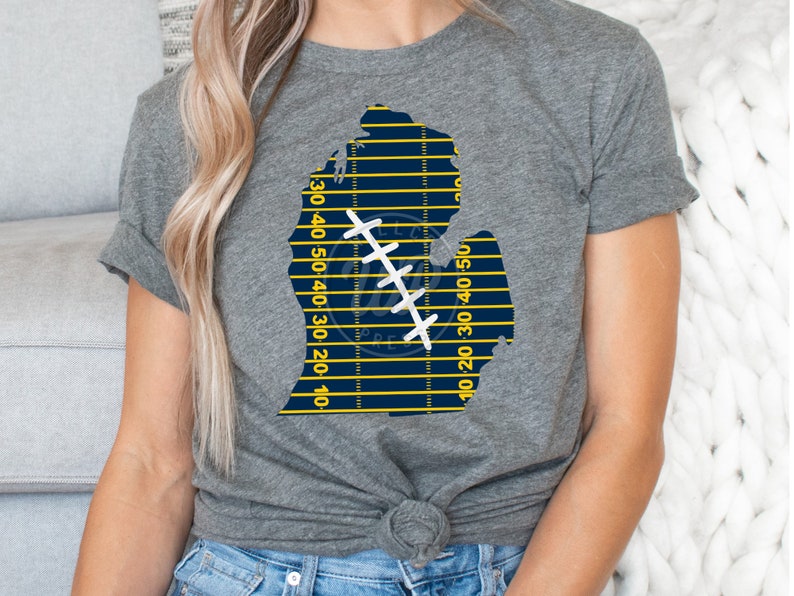 May include: A gray t-shirt with a blue and yellow outline of the state of Michigan. The outline is filled with a football field pattern and a white stitched football.