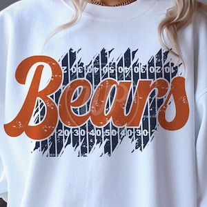 May include: White sweatshirt with a vintage-style orange graphic that says "Bears" on a blue and white striped background. The stripes have numbers on them.