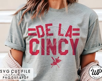 Può includere: Una maglietta grigia con il testo "DE LA CINCY" in rosso. Il testo è in grassetto, con un carattere a blocchi. C'è un piccolo grafico di una mazza da baseball e una scarpa con le ali sotto il testo.
