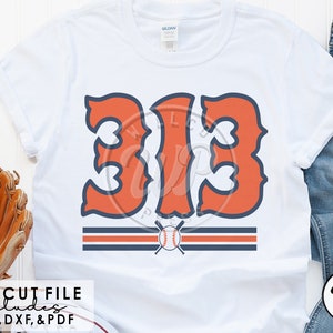 Puede incluir: Una camiseta blanca con un diseño de número de béisbol "313" rojo y azul. El número está en una fuente de estilo vintage con una pelota de béisbol y bates cruzados debajo. El diseño es perfecto para un fanático del béisbol.