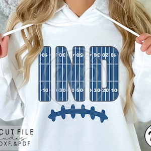 Puede incluir: Sudadera con capucha blanca con la palabra "IND" en azul, diseñada para parecerse a un campo de fútbol. Debajo de las letras hay una forma de balón de fútbol azul. La sudadera con capucha también presenta el texto "SVG CUT FILE includes PNG, DXF, & PDF".