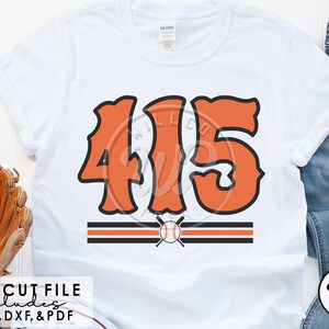 Op de afbeelding: Een wit T-shirt met het nummer "415" in oranje in een retro-baseballlettertype. Het nummer is zwart omrand en heeft twee zwarte strepen met een honkbal en gekruiste knuppels ertussen.