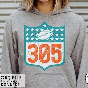Puede incluir: Una sudadera gris con un diseño desgastado en turquesa y naranja que presenta un balón de fútbol y el número 305. El diseño tiene la forma de un escudo con estrellas alrededor de los bordes.