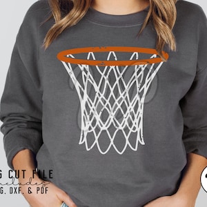 May include: A white basketball hoop with an orange rim on a gray sweatshirt. The hoop has a distressed look. The text "SVG CUT FILE includes PNG, DXF, & PDF" is at the bottom of the image.