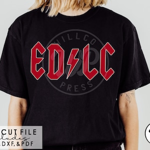 以下が含まれることがあります： 黒いTシャツに、赤い「ED/LC」の文字が太字でスタイリッシュなフォントで描かれています。文字は稲妻で囲まれています。「WILLCO PRESS」の文字がグラフィックの下に見えます。