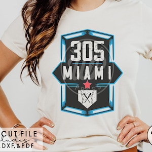 Puede incluir: Una camiseta blanca con un gráfico de béisbol azul y negro que dice "305 Miami". El gráfico tiene una estrella roja y una pelota de béisbol con bates cruzados. El texto "SVG CUT FILE includes PNG, DXF, & PDF" está en la parte inferior de la imagen.