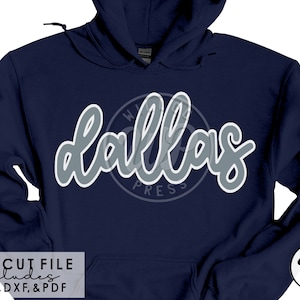 Op de afbeelding: Een marineblauwe hoodie met een witte en grijze "Dallas" grafische afbeelding op de voorkant. De afbeelding is in een schrijflettertype en heeft een witte omtrek. De hoodie heeft een capuchon met trekkoord en een kangoeroezak.