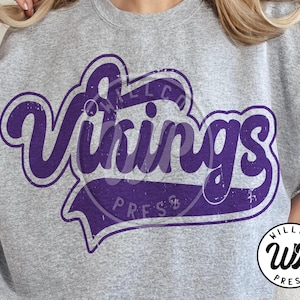 Può includere: Una felpa grigio melange con la scritta "Vikings" in viola invecchiato. Il design include uno stendardo con la parola "Press" e un logo circolare con la scritta "Willco Press".