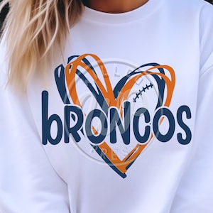 Puede incluir: Sudadera blanca con un diseño de corazón azul y naranja y la palabra "broncos" en letras azules.