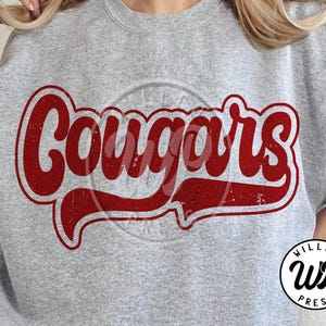 Pode incluir: Um moletom cinza com a palavra "Cougars" em letras vermelhas desgastadas. O texto tem um estilo vintage e retrô. O moletom também apresenta um pequeno logotipo circular com o texto "WILLCO PRESS".