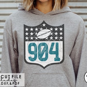 May include: A teal blue and white distressed design with the number 904 and a football inside a shield shape with stars. The design is on a gray hoodie.