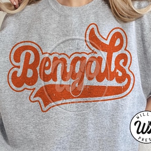 May include: A heather gray sweatshirt featuring the word "Bengals" in a distressed orange font. The text is in a retro style with a shadow effect. A circular logo with the text "WILLCO PRESS" is in the bottom right corner.