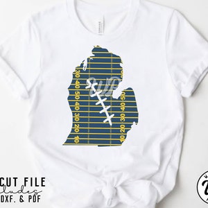 May include: A white t-shirt with a blue and yellow Michigan state outline design. The outline is shaped like a football field with yard lines and a football player graphic. The text "SVG CUT FILE includes PNG, DXF, & PDF" is at the bottom of the image.