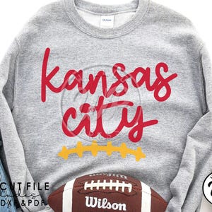 May include: Gray sweatshirt with the text "Kansas City" in red lettering. The text is in a cursive font. There are five yellow stars underneath the text. A brown and white football with the word "Wilson" on it is in the bottom right corner of the image.