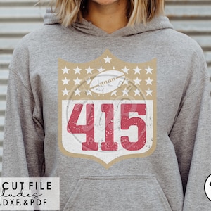 Op de afbeelding: Een grijze hoodie met een vintage-stijl football schildontwerp met het nummer 415 in rood met een witte omtrek. Het schild is omringd door gouden sterren en een witte voetbal.