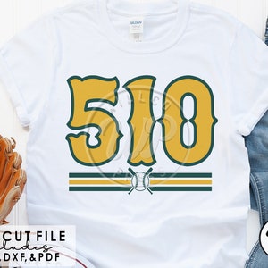 Könnte beinhalten: Ein weißes T-Shirt mit einem grünen und goldenen Baseball-Design. Das Design zeigt die Nummer "510" in einer Retro-Schriftart mit einem Baseball und gekreuzten Schlägern darunter.