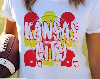 May include: White t-shirt with a red, yellow, and white graphic design that says "Kansas City". The graphic design features football helmets.