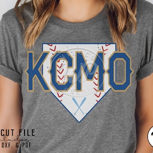 Può includere: Una maglietta grigia con un design a forma di diamante da baseball blu e oro con il testo "KCMO". Il diamante è riempito con cuciture da baseball bianche e rosse.