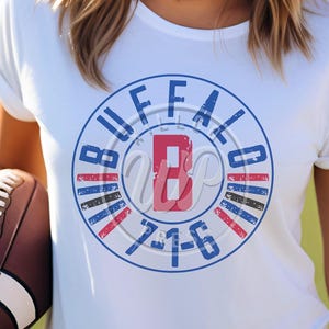Op de afbeelding: Wit T-shirt met een blauwe en rode cirkelvormige afbeelding met de tekst "Buffalo" met een rode "B" in de cirkel en de tekst "7-1-6" onder de "B".