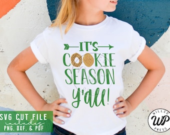 May include: A white t-shirt with green text that reads "It's Cookie Season y'all!" with a brown cookie graphic. The shirt is being worn by a child.