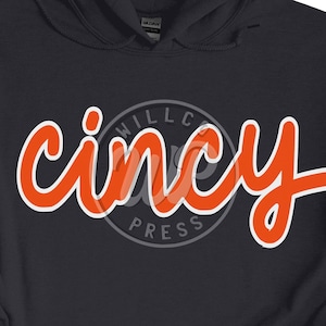 Puede incluir: Una sudadera con capucha azul oscuro con un gráfico blanco y naranja que dice "cincy" con un círculo alrededor que dice "Willco Press".