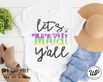 Könnte beinhalten: Weißes T-Shirt mit dem Text "Let's Mardi Y'all" in schwarzer und violetter Schrift mit grünen Akzenten. Das Shirt liegt auf einer weißen Holzoberfläche mit einem braunen Hut, blauen Jeans und braunen Schuhen.