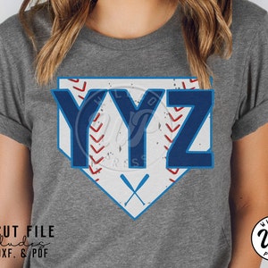 May include: Gray t-shirt featuring a baseball-themed design. The design includes the letters "YYZ" in blue, set against a white and blue home plate shape, with red baseball stitching and crossed bats. The shirt also includes the text "SVG CUT FILE includes PNG. DXF. & PDF."