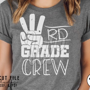 May include: A gray t-shirt with white text that reads "3rd Grade Crew" with a hand making the number three sign.