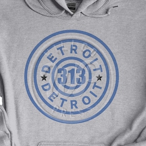 Puede incluir: Una sudadera gris con un diseño circular azul que presenta el texto "DETROIT" y el número "313" en el centro.