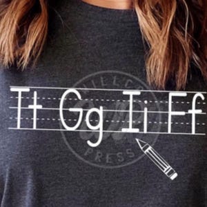 May include: A dark gray t-shirt with white text that says "Tt Gg Ii Ff" with a pencil drawn underneath. The text is written on a lined sheet of paper.