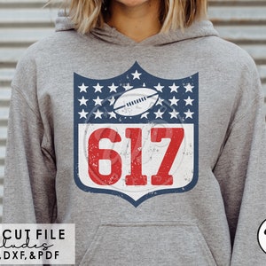 May include: A gray hoodie with a blue and red distressed shield design featuring a football and the number 617. The shield is surrounded by white stars on a blue background.