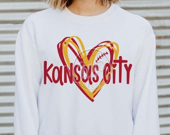 May include: White sweatshirt with a red heart design and the text "Kansas City" in red. The heart has a yellow outline and a baseball inside.