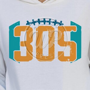 Puede incluir: Sudadera blanca con un diseño en turquesa y naranja que presenta el número "305" con un gráfico de fútbol arriba.