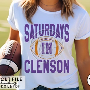 May include: A white t-shirt with purple text that reads "Saturdays in Clemson" and a purple and orange football graphic. The text "SVG Cut File Includes PNG, DXF, & PDF" is at the bottom of the image.