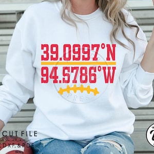 May include: White sweatshirt with red and yellow text that reads "39.0997°N 94.5786°W" and a yellow football graphic. The text "PRES" is below the graphic.